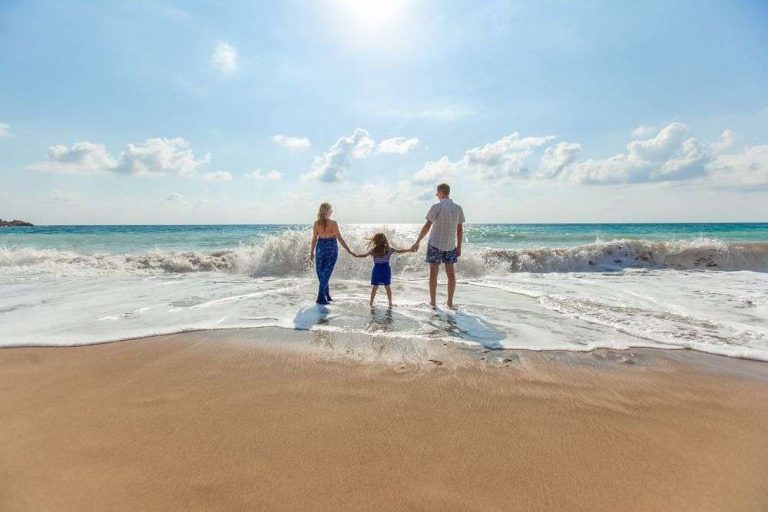 A family stands hand-in-hand at the shoreline, facing the ocean waves under a bright sky.