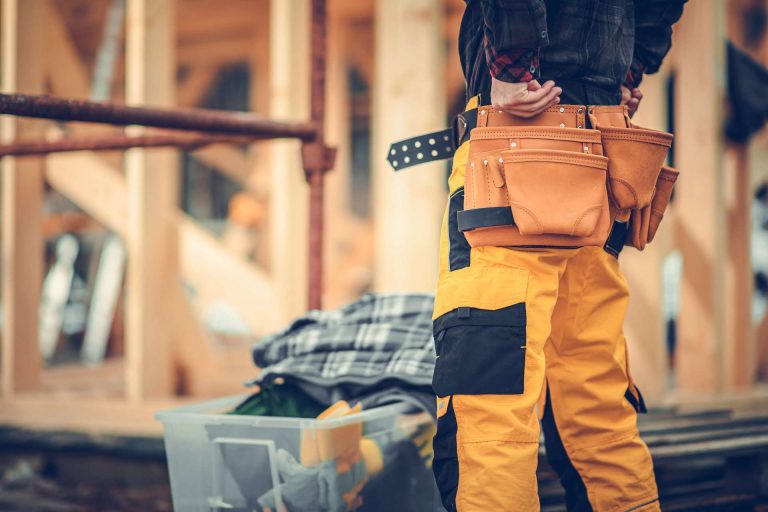 A worker in yellow pants and a tool belt stands in a construction area with wooden framing in the background.