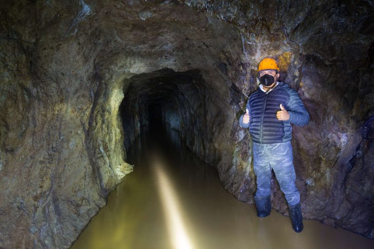 A person wearing a helmet and mask stands in a dimly lit underground tunnel with water on the ground.