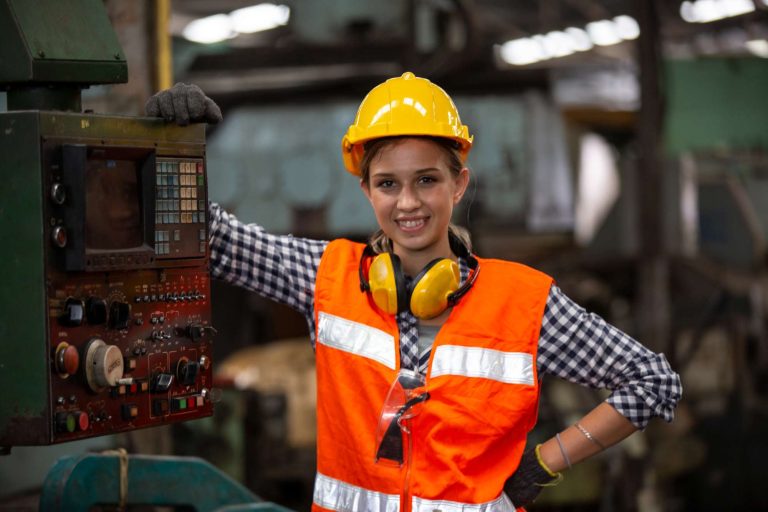 A woman in a hard hat and safety vest stands next to industrial machinery, smiling confidently.