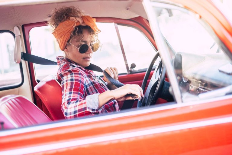 A woman with curly hair and sunglasses fastens her seatbelt in a vintage red car.