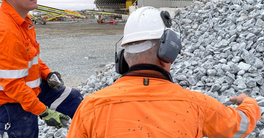 Two workers in orange safety gear discuss near a pile of gravel at a construction site.