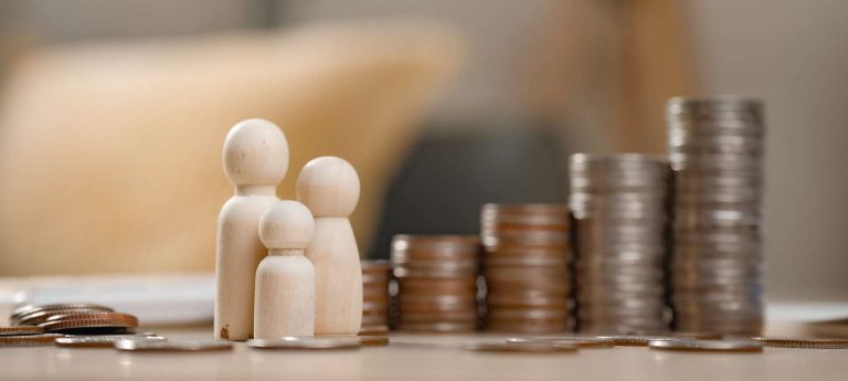 Wooden figurines of a family stand in front of stacks of coins on a table.