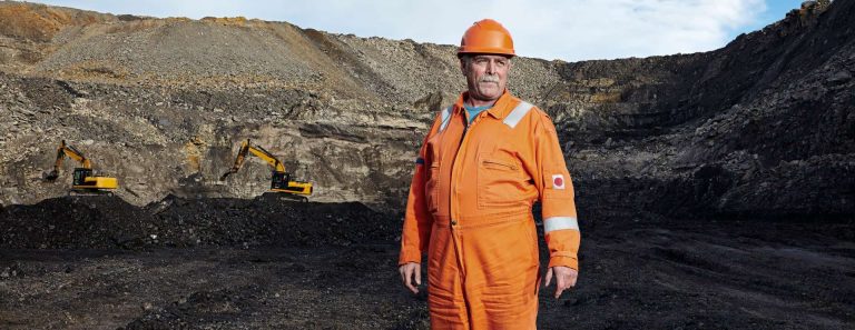 A worker in an orange safety suit stands in a mining site with excavators in the background.