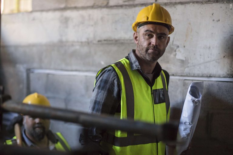 A construction worker in a hard hat and reflective vest holds blueprints while standing in a building under construction.