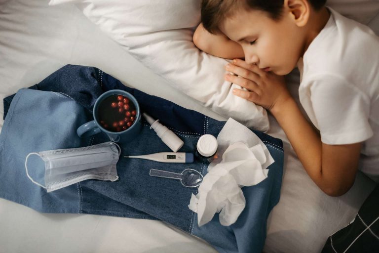 A young boy sleeps on a bed next to a cup of berries, a thermometer, a mask, and other health items.