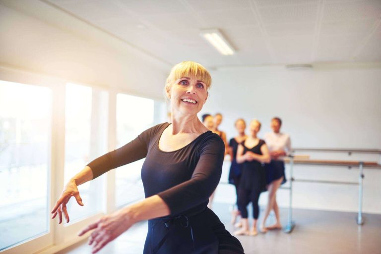 A smiling ballet instructor demonstrates a dance move in a studio with students in the background.