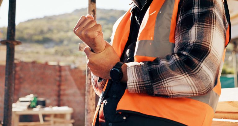 A construction worker in a safety vest adjusts their wrist while standing at a worksite.