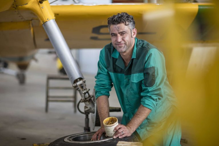 A mechanic in a green jumpsuit smiles while working on an aircraft tire in a hangar.