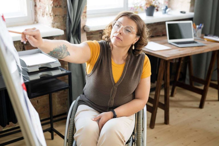 A woman in a wheelchair points at a whiteboard while seated in a modern office space.