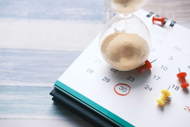 An hourglass sits on a calendar marked with the date 30, surrounded by colorful push pins.