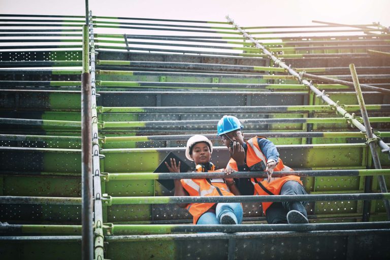 Two construction workers in safety gear sit on scaffolding, reviewing plans and discussing their work.