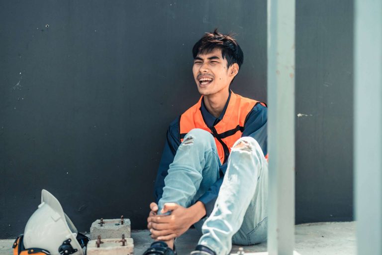A young man in an orange safety vest sits on the ground, smiling and resting against a wall.