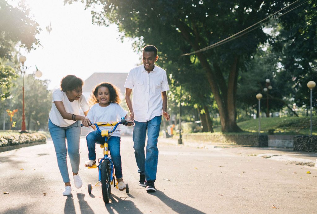 A child rides a bicycle while being assisted by two adults in a sunny park setting.
