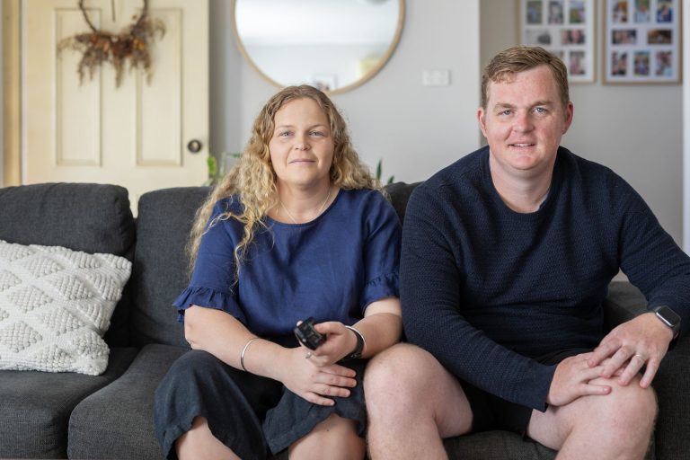 A couple sits together on a couch in a cozy living room, smiling at the camera.