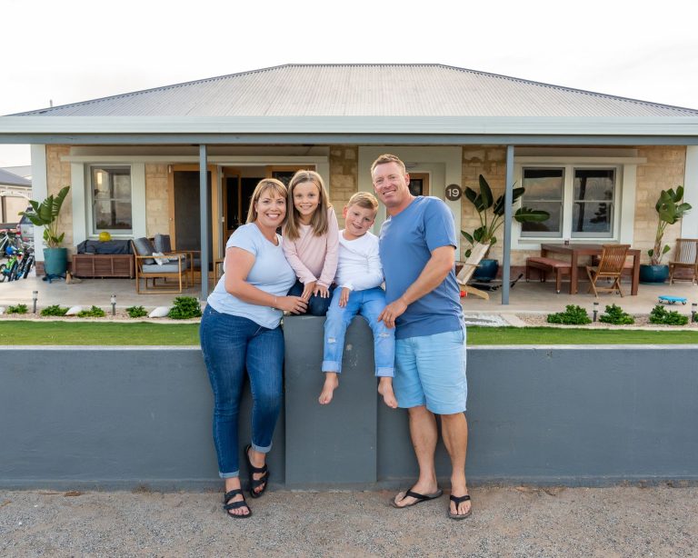A family of four poses together in front of their home, smiling and enjoying the moment.