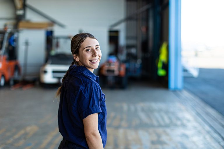 A smiling woman in a blue work shirt stands in a warehouse with vehicles in the background.