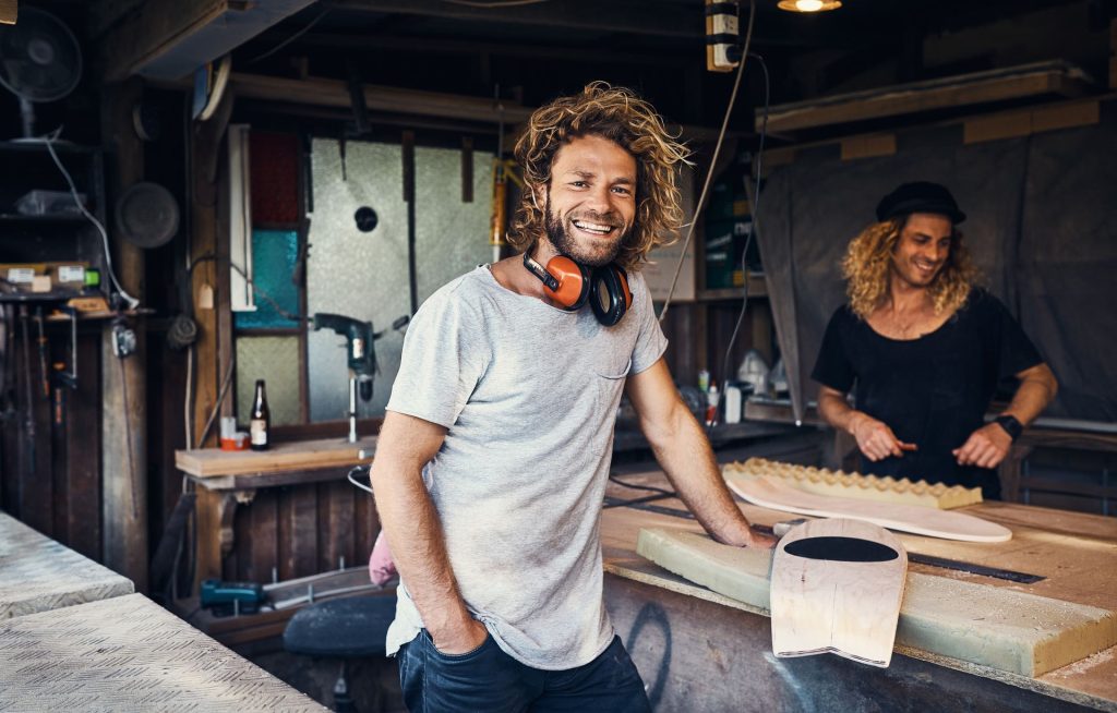 Two men working in a workshop, one smiling while leaning on a workbench with tools and materials around them.