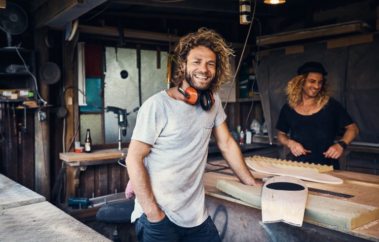 Two men working in a workshop, one smiling while leaning on a workbench with tools and materials around them.