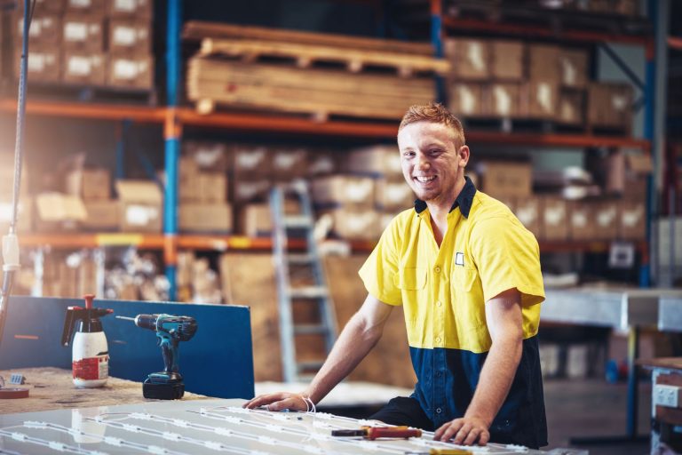 A smiling worker in a yellow and blue shirt stands at a table with tools in a warehouse setting.