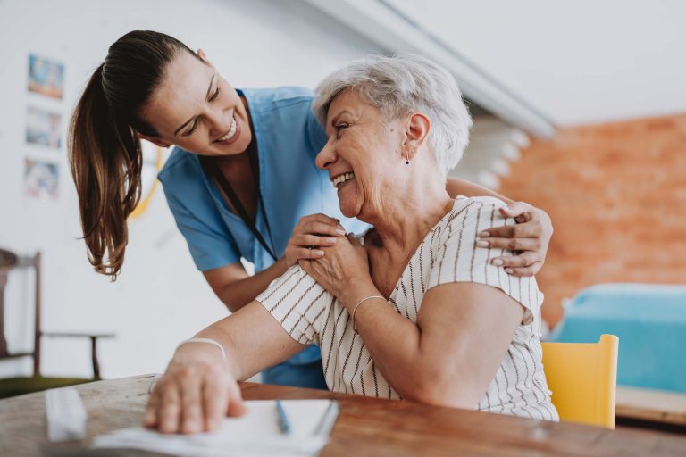 A caregiver smiles and comforts an elderly woman during a friendly interaction at home.