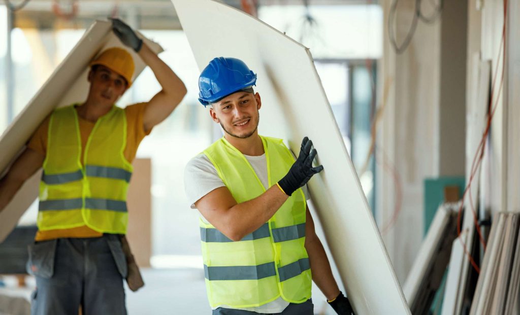 Two construction workers in safety vests and hard hats carry drywall sheets in a partially finished building.
