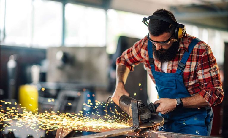 A man in protective gear operates a grinder, creating sparks in a workshop.