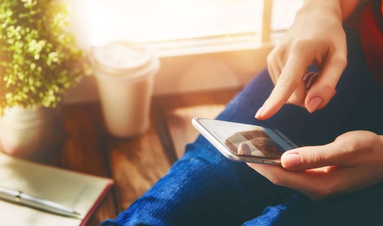 A person using a smartphone while sitting next to a small plant and a coffee cup.