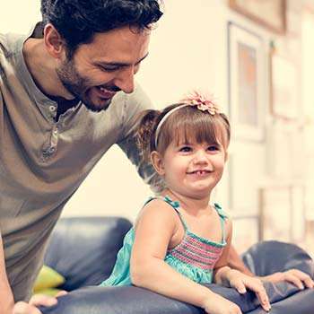 A smiling father interacts with his young daughter in a cozy living room setting.
