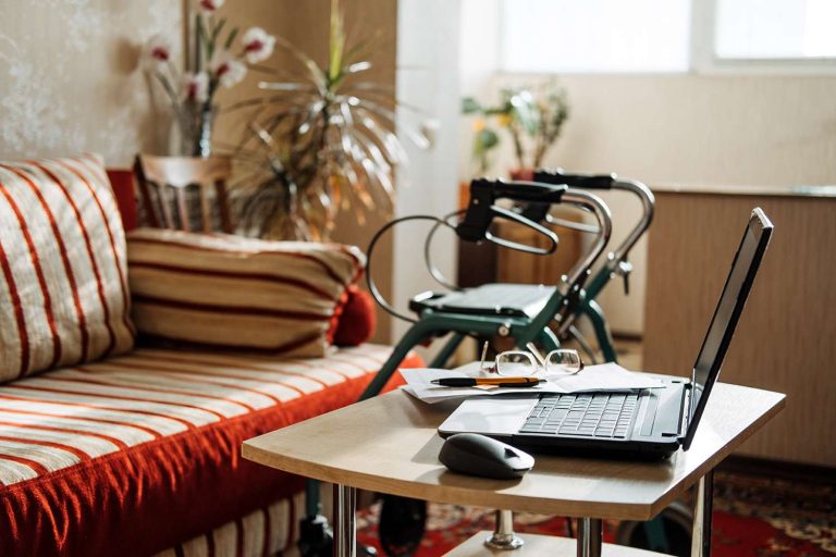 A laptop, glasses, and papers are on a table beside a striped couch and a walker in a cozy living room.