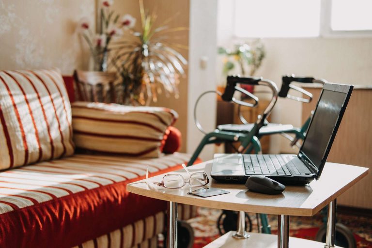 A laptop and glasses are on a small table next to a striped sofa and a walker in a bright living space.