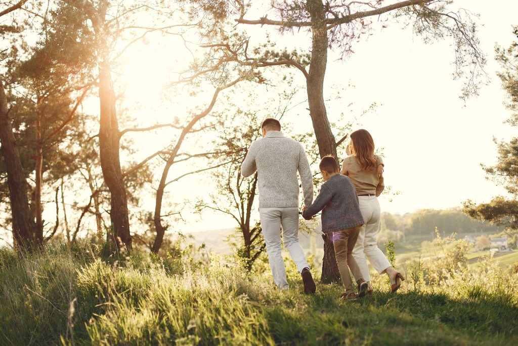 A mum, dad and young son walking hand in hand through sunlit trees at golden hour.