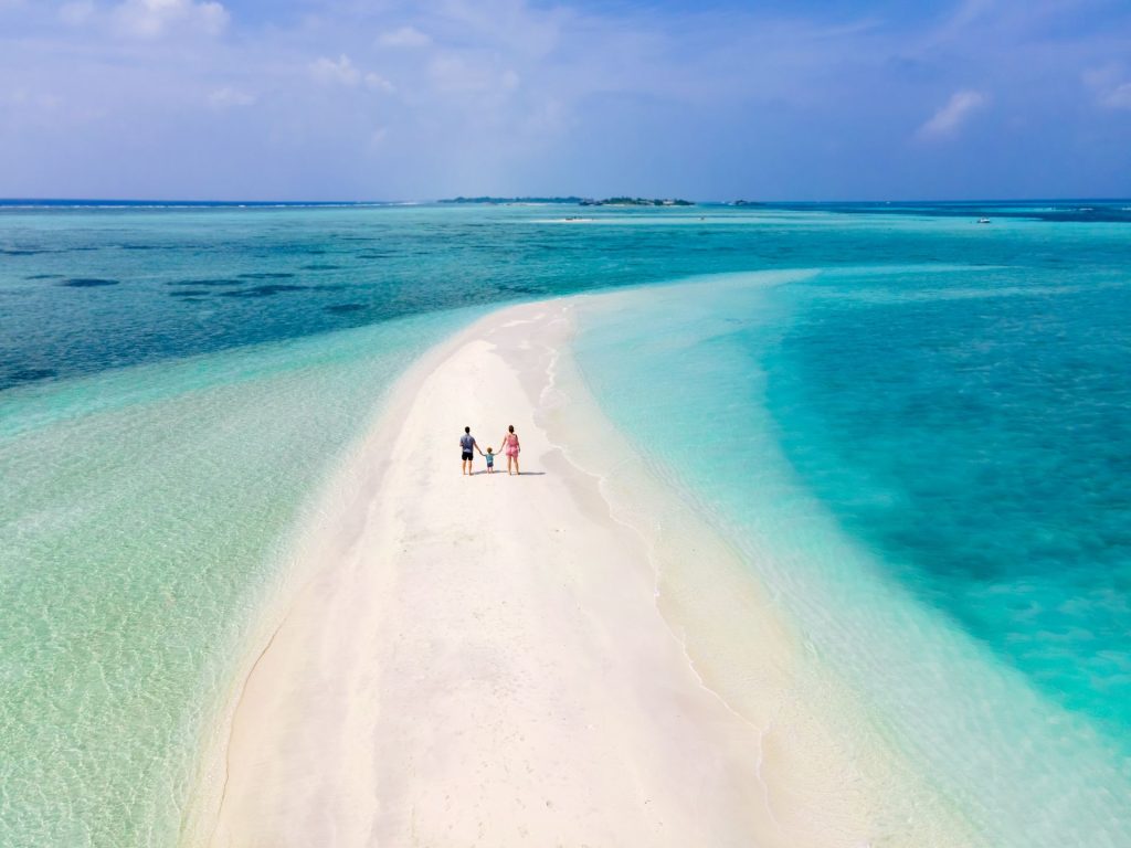 A family walks hand in hand along a sandy beach with clear turquoise waters.