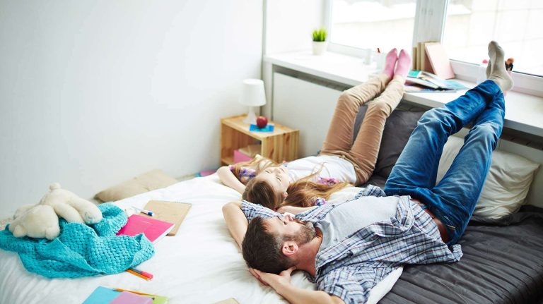 A couple relaxes on a bed with their feet up, surrounded by books and a teddy bear.