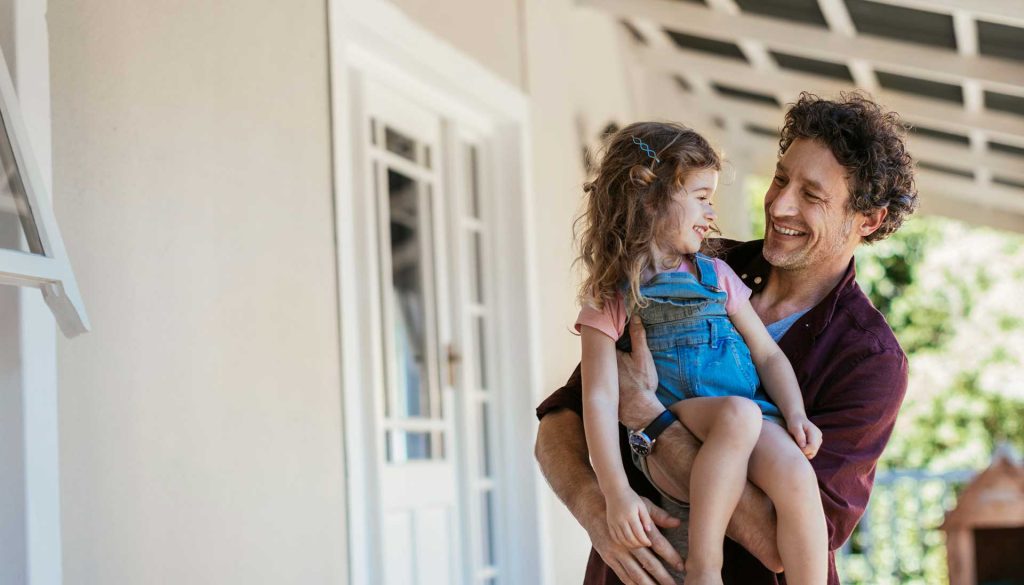 A smiling man holds a young girl in overalls, both enjoying a moment together on a porch.