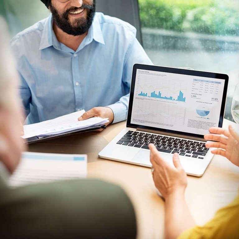 A smiling man discusses data displayed on a laptop with two other people at a table.