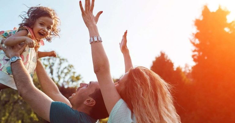 A joyful family enjoys a sunny day outdoors, with a parent lifting a smiling child into the air.
