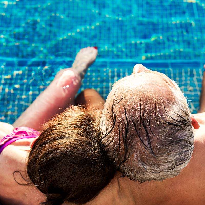 An older couple relaxes together by a swimming pool, with water glistening in the background.