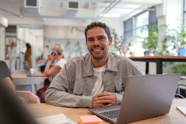 Smiling man sitting at a desk with a laptop in a bright, modern office space.