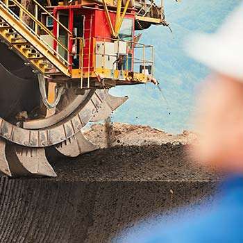 A large mining machine with a rotating cutter wheel is excavating earth, with a blurred figure in the foreground.