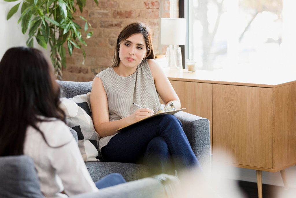A woman takes notes while listening to another person in a cozy office setting.