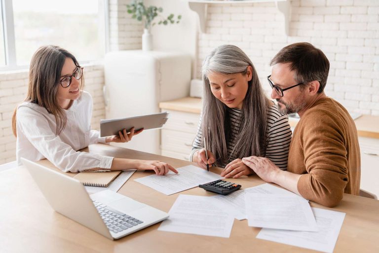 A woman assists a couple with paperwork and calculations at a table, with a laptop and documents visible.