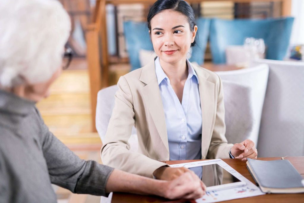 A professional woman in a suit engages with an elderly client at a table, discussing documents and a tablet.