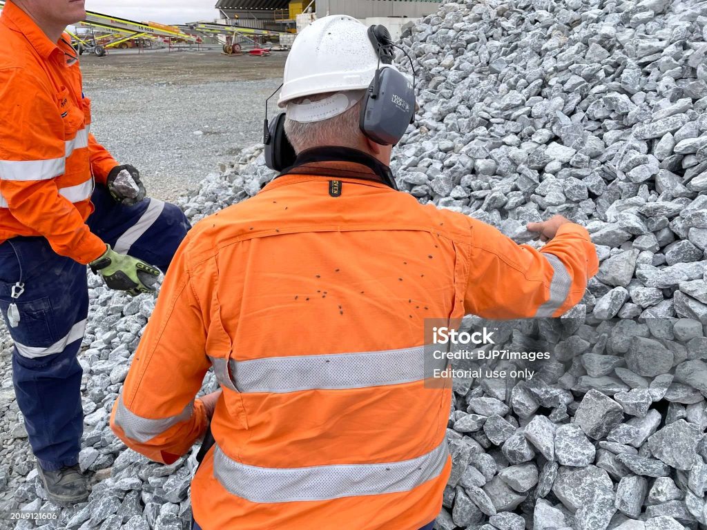 Two workers in orange safety gear are examining a pile of gravel at a construction site.