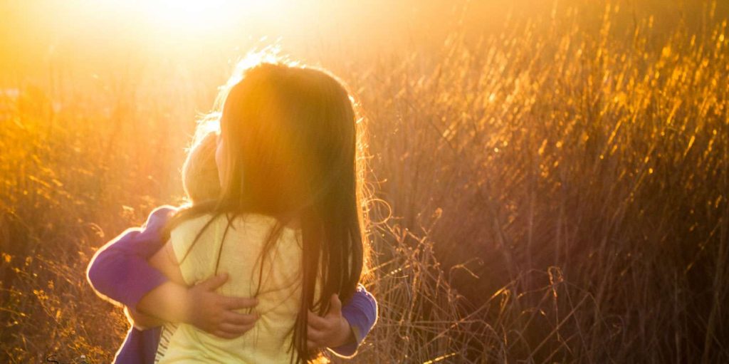 Two children embrace in a sunlit field with tall grass.