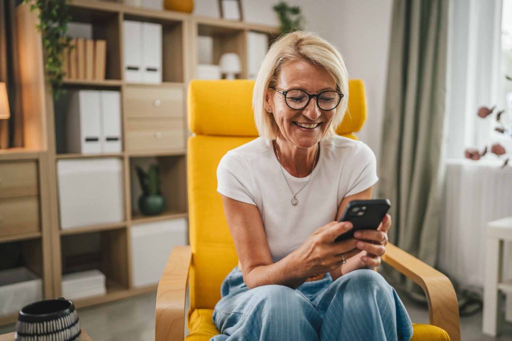 A smiling woman in glasses sits in a yellow chair, looking at her smartphone in a cozy, modern living space.