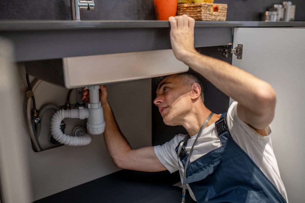 A person inspects plumbing under a kitchen sink, focusing on the pipes and connections.