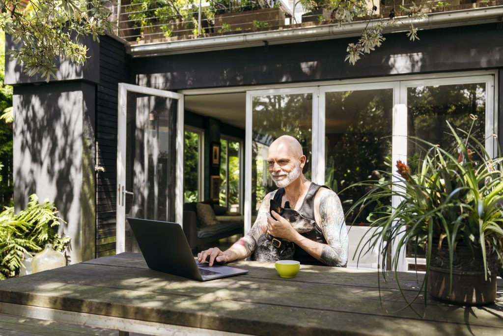 Man with tattoos sitting at a table outdoors, using a laptop and holding a small dog.