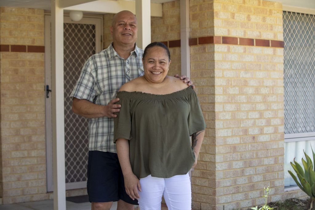A couple stands together smiling in front of a brick house.