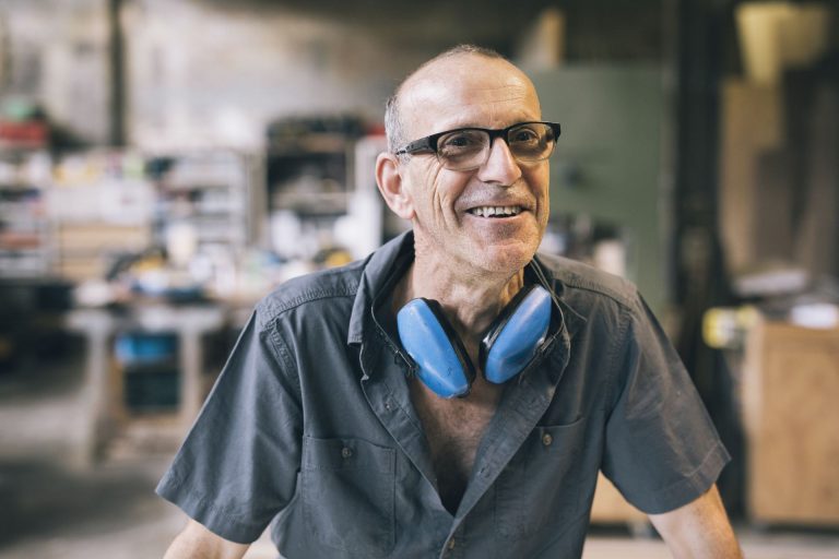 Smiling older man wearing glasses and ear protection in a workshop setting.